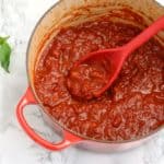 marinara sauce in a red pot with red cooking spoon inserted in the pot.