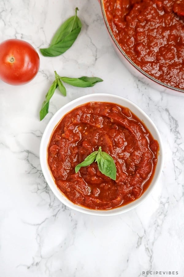 Marinara sauce in a white bowl garnished with basil with some part of the pot of marinara in view.