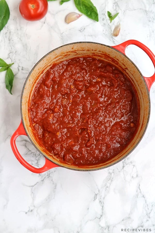 Marinara sauce in a red pot with basil, garlic and one tomato on the table beside the pot.