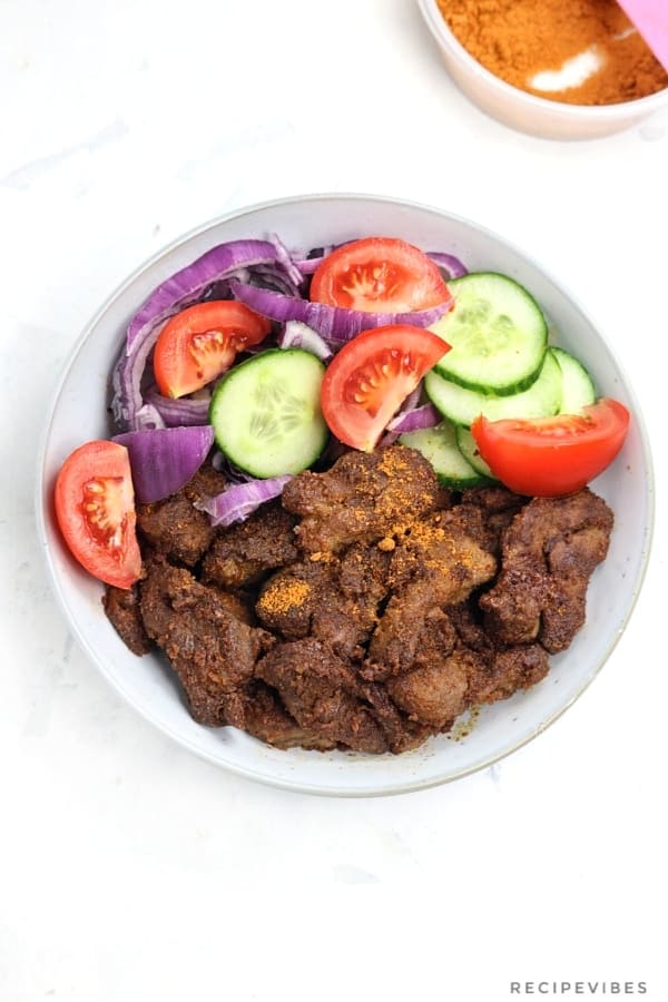 Beef suya sprinkled with yaji served in a plate and garnished with onion, tomatoes and cucumber slices.